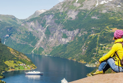 Frau sitzt auf einem Felsen und schaut auf den Geiranger Fjord hinunter